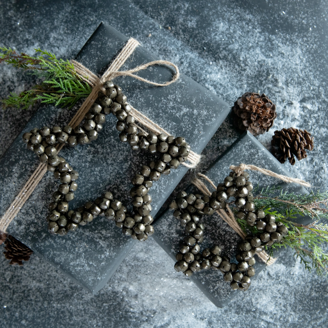 Jingle bell star shaped ornament tied onto a decorative gift with pine cones and greenery on a textured surface