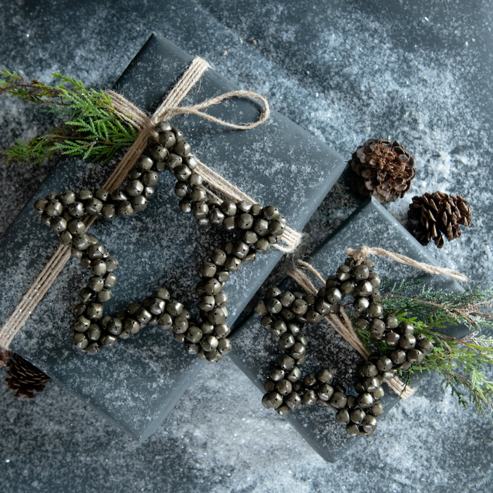 Jingle bell star shaped ornament tied onto a decorative gift with pine cones and greenery on a textured surface