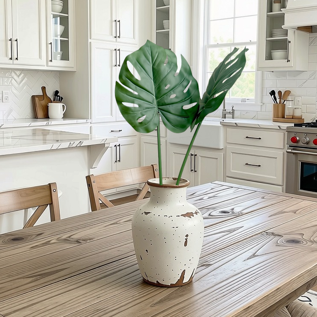 A rustic white ceramic vase with distressed brown speckles sits on a light wood dining table in a bright, modern kitchen. The vase holds two large green monstera leaves, which serve as the focal point of the composition.