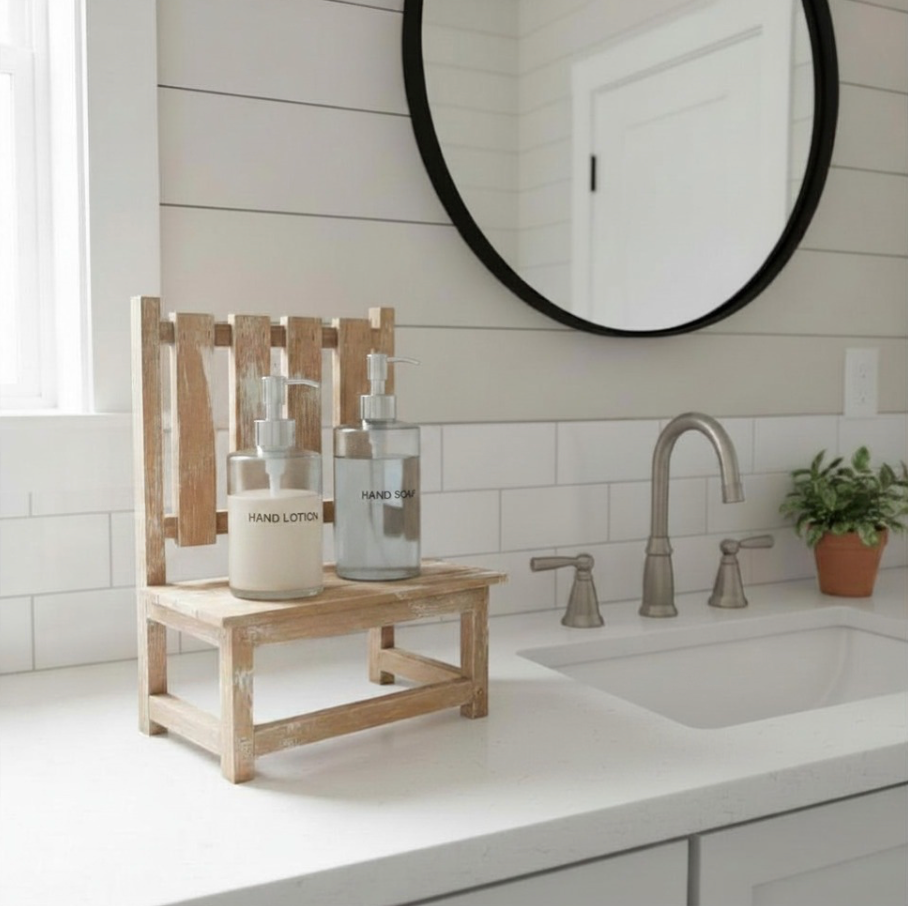 A small rustic wooden bench stand holding two soap dispenser bottles on a bathroom counter, with a white sink, tiled wall, round mirror, and a small potted plant nearby.