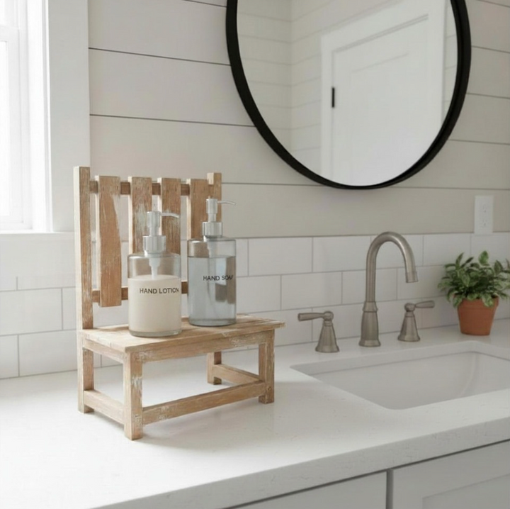 A small rustic wooden bench stand holding two soap dispenser bottles on a bathroom counter, with a white sink, tiled wall, round mirror, and a small potted plant nearby.