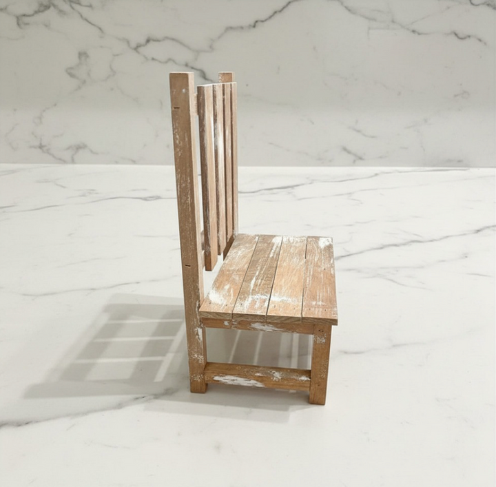 A miniature wooden chair with a distressed, whitewashed finish, shown from a side angle on a white marble surface with a matching marble background.
