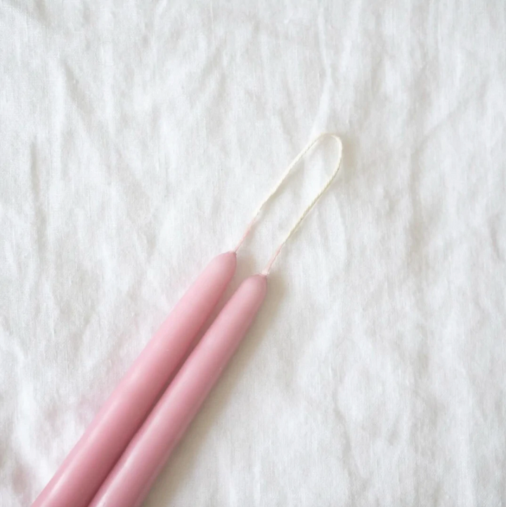 A selection of 12-inch taper candles in beige, pink, black, and gray, displayed hanging on a wooden rod against a neutral background.