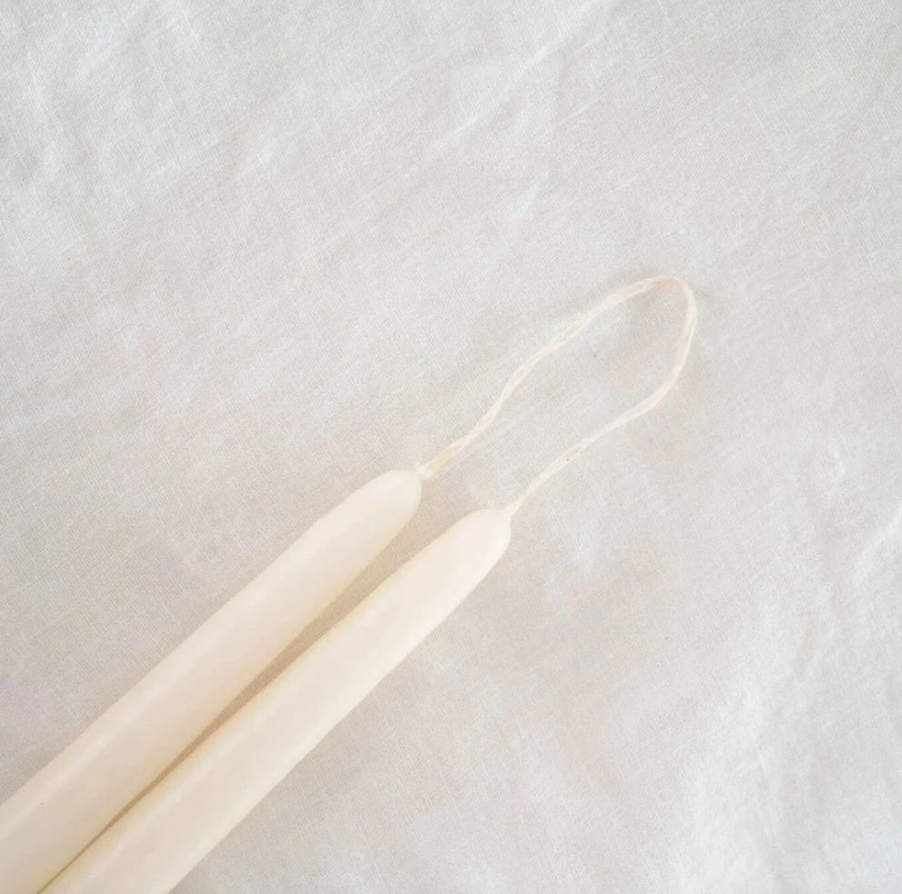 A selection of 12-inch taper candles in beige, pink, black, and gray, displayed hanging on a wooden rod against a neutral background.