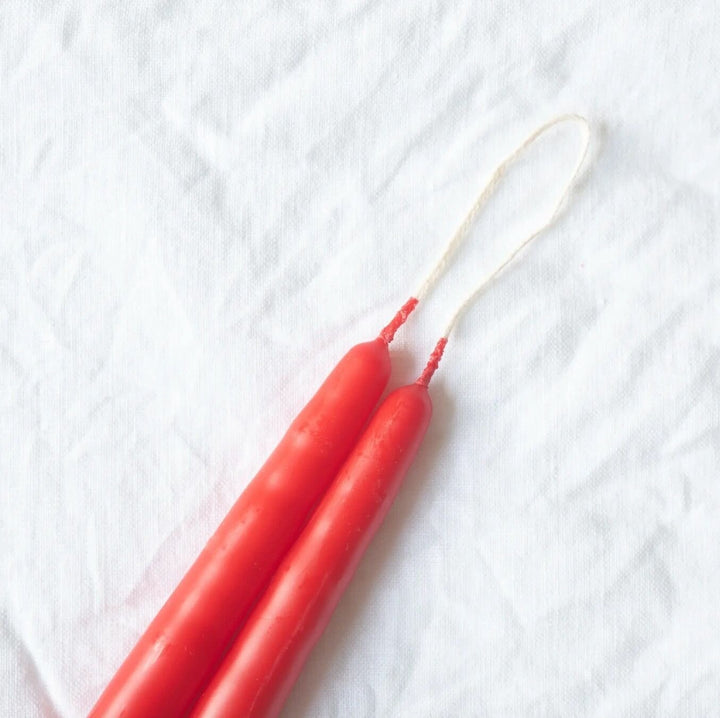 A selection of 12-inch taper candles in beige, pink, black, and gray, displayed hanging on a wooden rod against a neutral background.