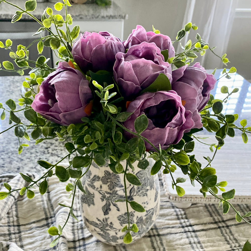 A bundle of artificial purple peony flowers with ruffled petals and green leaves, tied with raffia, arranged in a ceramic vase with a floral pattern.