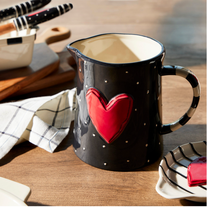 A black and white pitcher with a red heart design and white polka dots on a table with a checkered napkin and wooden cutlery in the background.