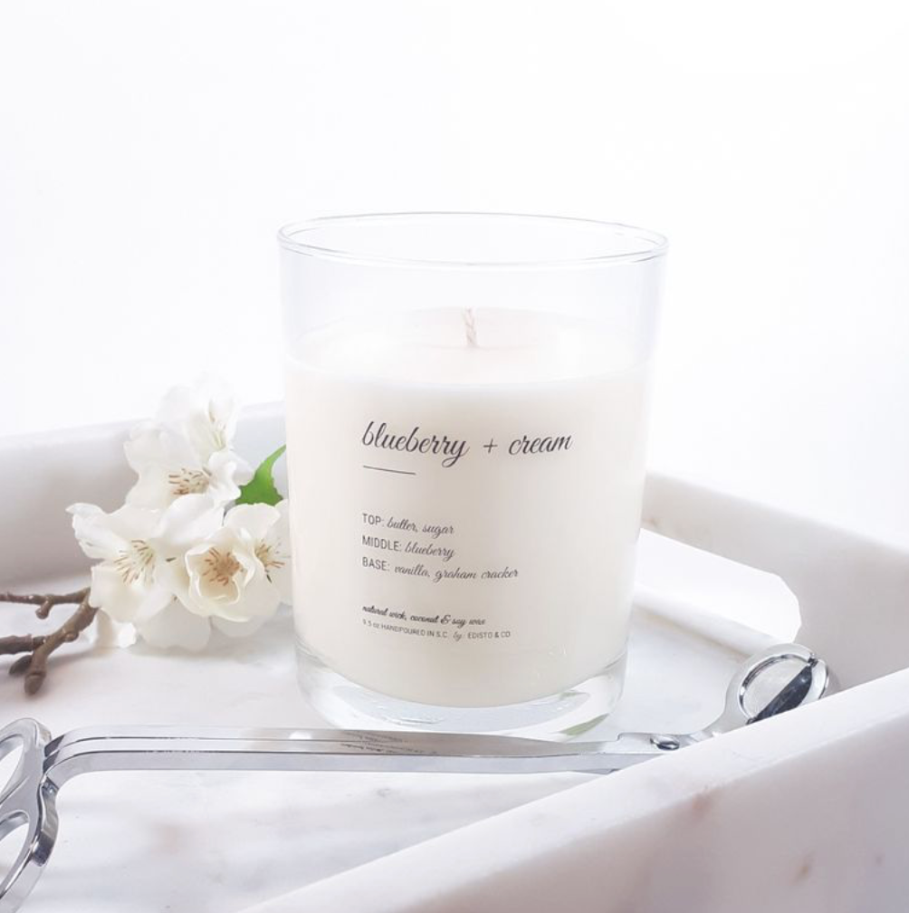 A white scented candle with a clear glass container, labeled 'Blueberry + Cream', placed on a white tray with a blurred background featuring white flowers.