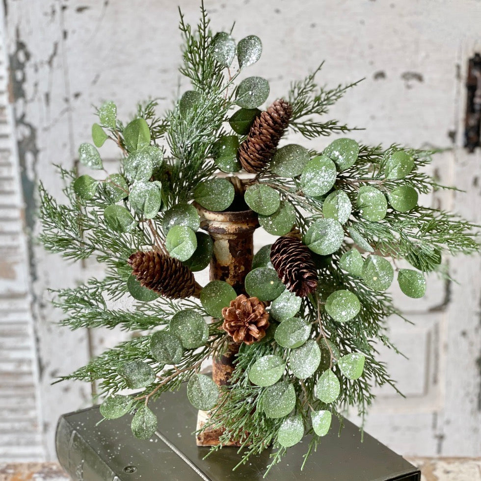 A decorative candle ring featuring green cedar, eucalyptus leaves, and pinecones, adorned with glitter and placed on a metal base.