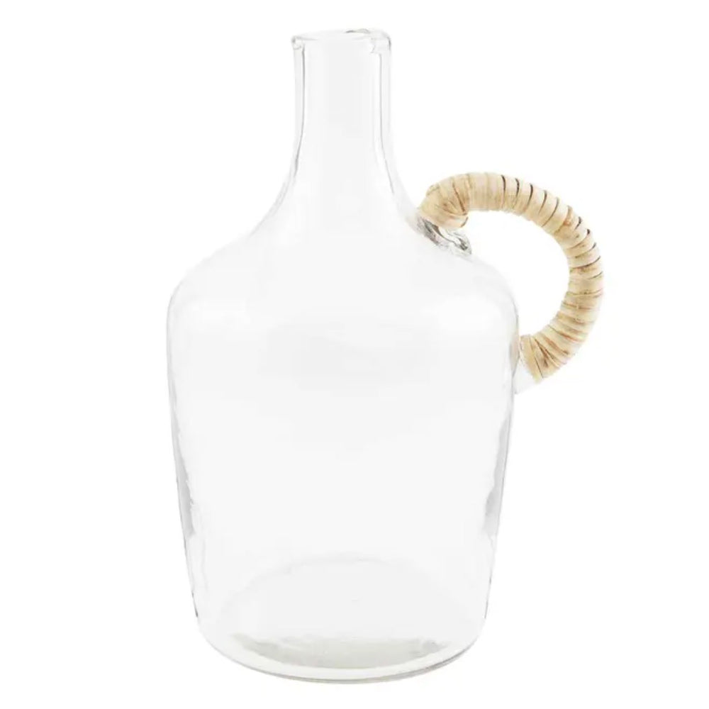 A clear glass jug with a wicker wrapped handle placed on a white surface, with a larger glass jug in the background containing some dried plants.