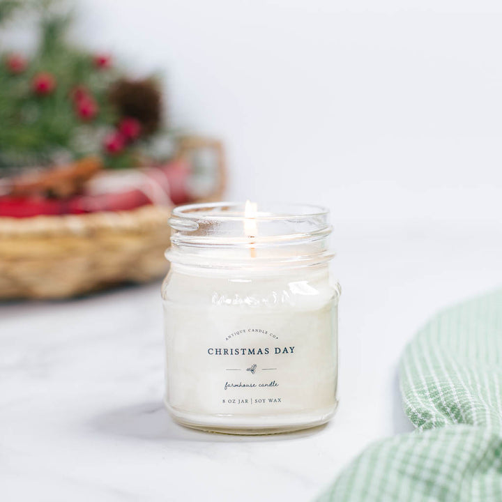 A scented candle in a clear mason jar with a reusable metal lid, labeled 'Christmas Day' and 'Farmhouse style', placed on a marble surface with a festive decor in the background.