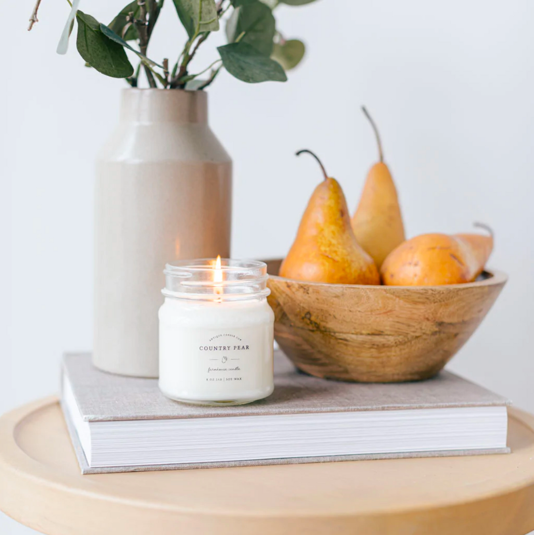 A lit Country Pear scented candle in a clear glass jar with a metal lid, placed on a book, with a vase of flowers and a bowl of pears in the background.