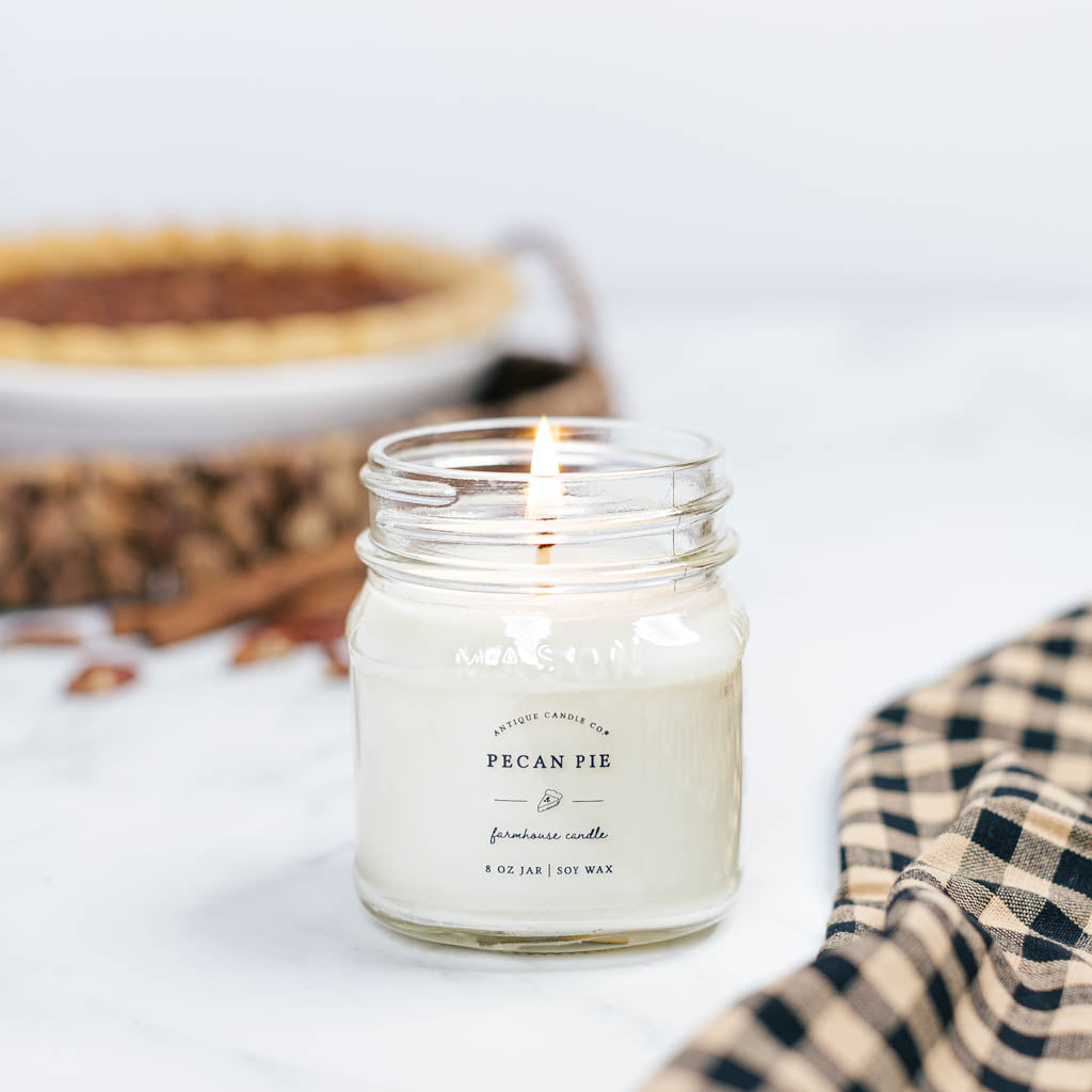 A PECAN PIE scented candle burning inside a clear glass jar with a metal lid, placed on a table with a checkered napkin and a pecan pie in the background.