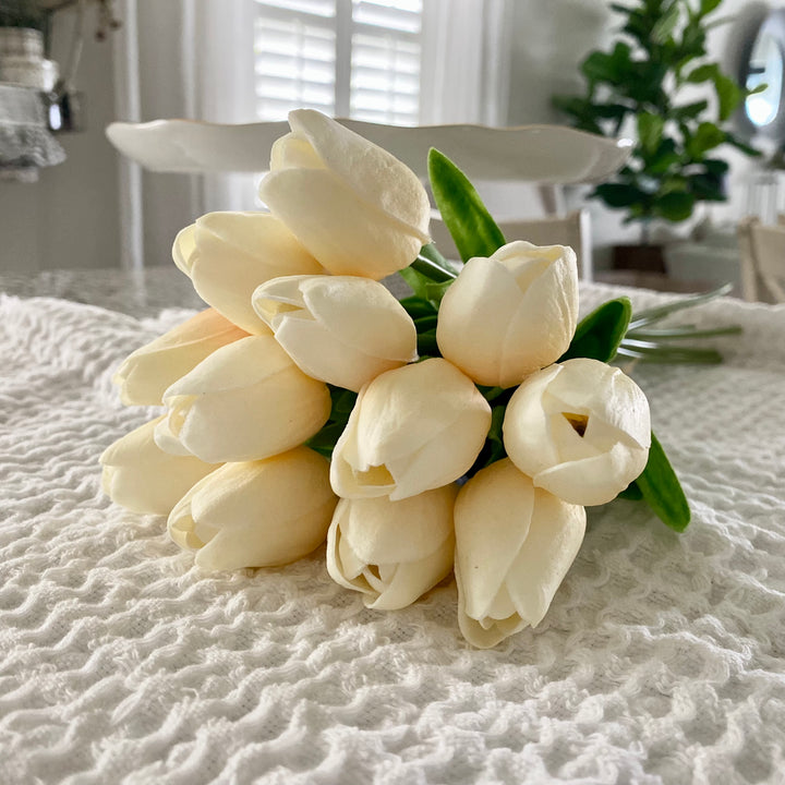 A bouquet of light peach, lifelike artificial tulips with green leaves, tied with a paper bow, placed on a textured surface.