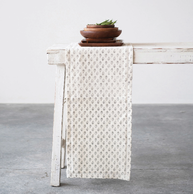 A floral pattern table runner in charcoal gray and cream colors, displayed on a white table with a wooden bowl on top.