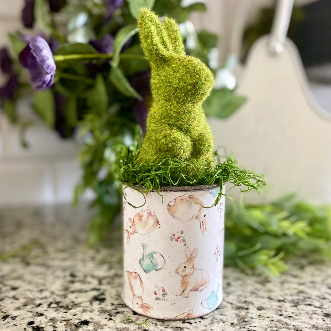 A galvanized metal container with a glossy paper label featuring a vintage design with a green bunny, a watering can, and strawberries on top, displayed on a kitchen counter.