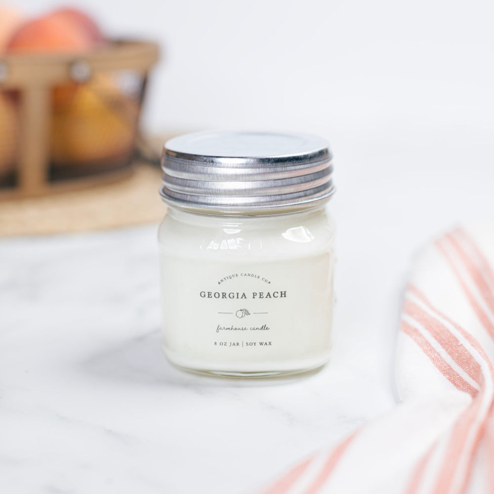 A Georgia Peach scented candle in a clear mason jar with a metal lid, placed on a table with a striped napkin and fruits in the background.
