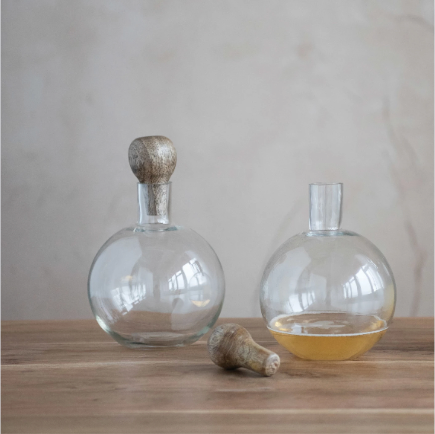 Clear glass decanter with mango wood stopper on a wooden surface against a neutral background.