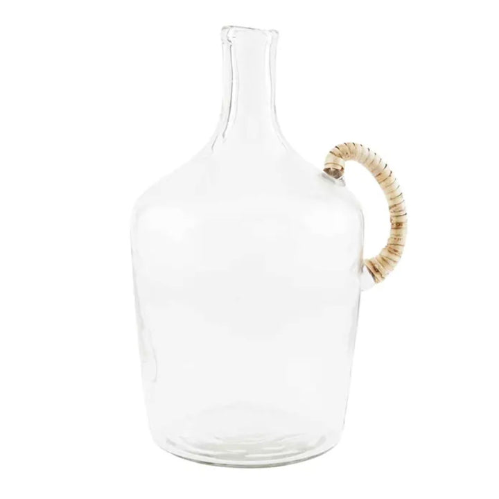 A clear glass jug with a wicker wrapped handle placed on a white surface, with a larger glass jug in the background containing some dried plants.