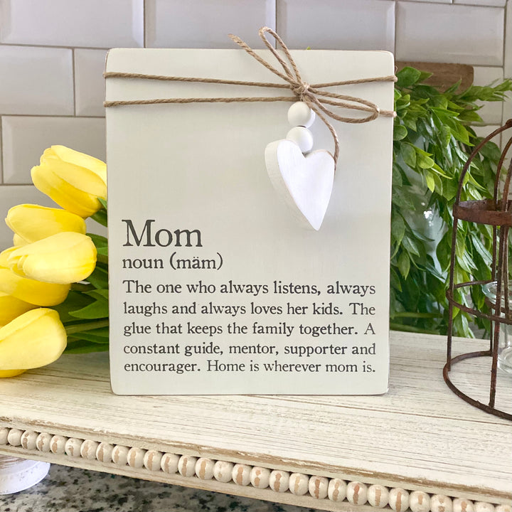 A large wooden plaque with 'Mom' written at the top, a white heart symbol, and the rest of the message that describes the characteristics of a mother. The plaque is twine-wrapped and set against a backdrop of yellow flowers and a metal bird feeder.