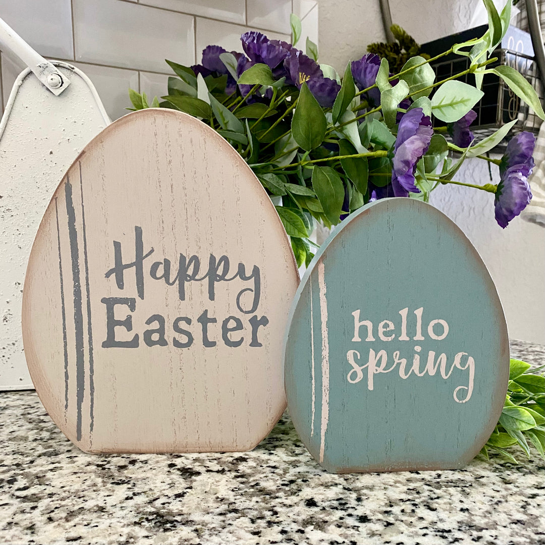 Two wooden egg-shaped decorations with the phrases 'Happy Easter' and 'Hello Spring' written on them, in shades of beige, teal, and gray, placed on a kitchen counter.