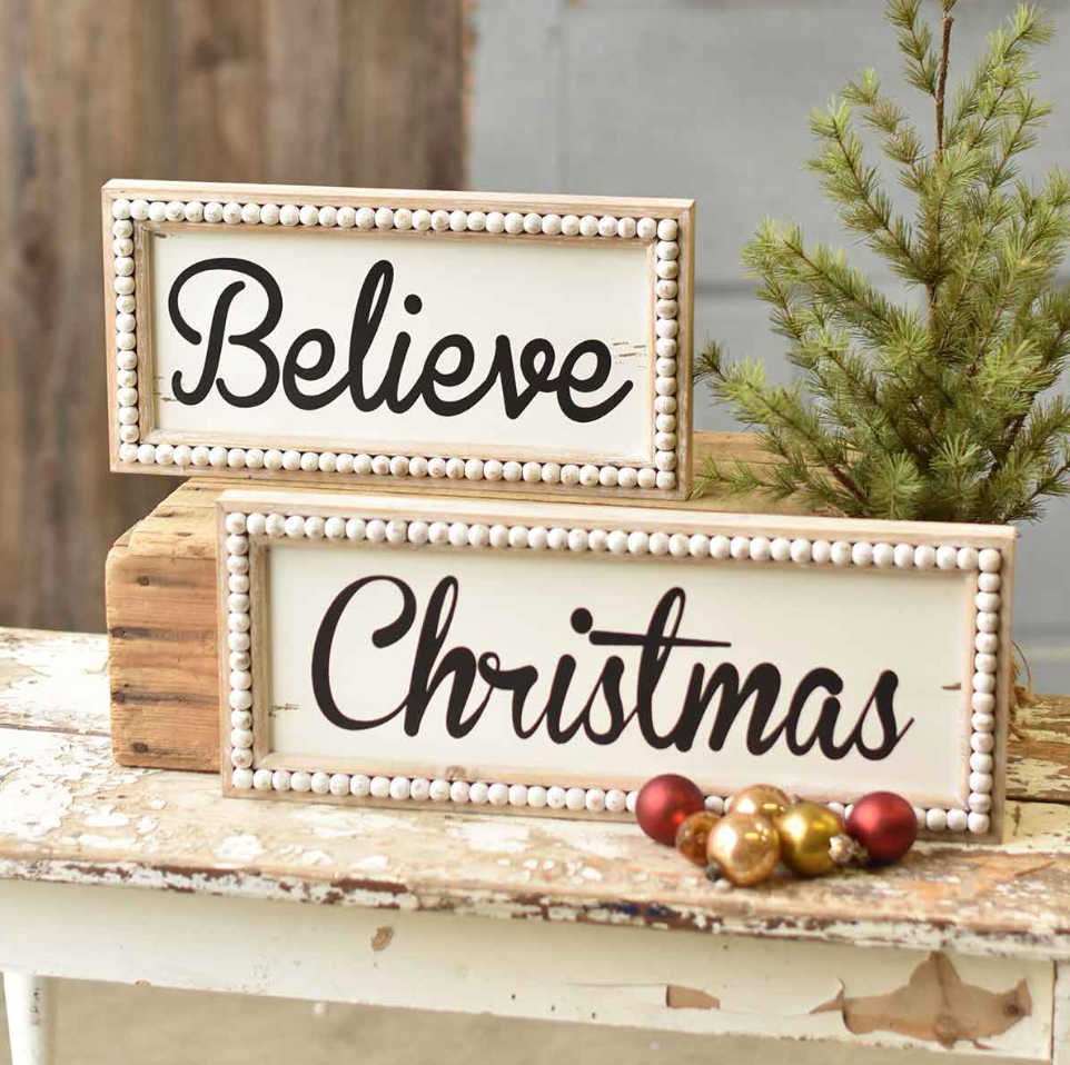 Two wooden beaded Christmas signs with the words 'BELIEVE' and 'CHRISTMAS' written in black script, placed on a rustic wooden surface with pine needles and holiday ornaments around.