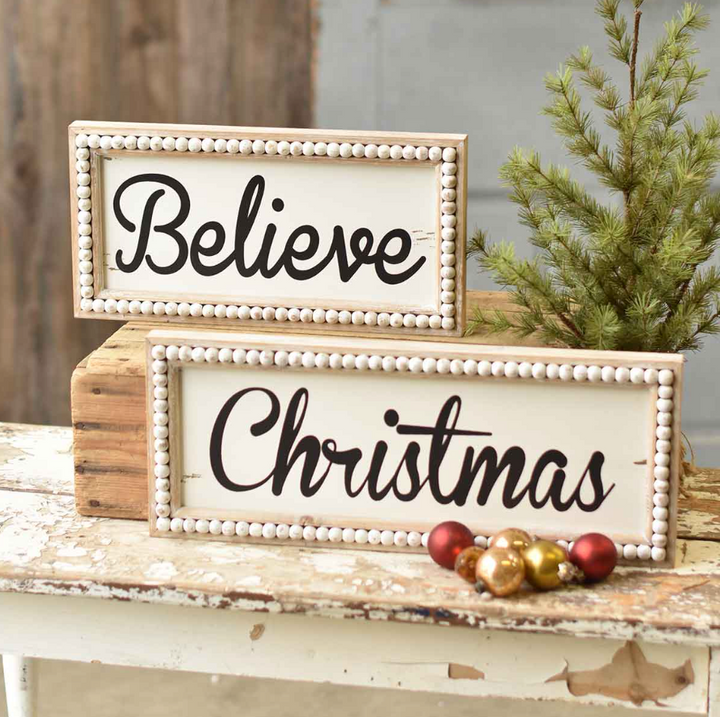 Two wooden beaded Christmas signs with the words 'BELIEVE' and 'CHRISTMAS' written in black script, placed on a rustic wooden surface with pine needles and holiday ornaments around.