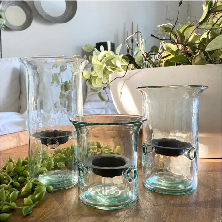 Three clear blown glass hurricane candleholders of varying sizes placed on a wooden surface with a plant in the background. Each holder has a removable metal candle cup.
