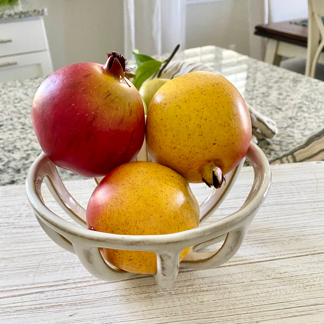 A set of three artificial pomegranates in red and yellow hues, displayed in a white bowl on a kitchen counter.
