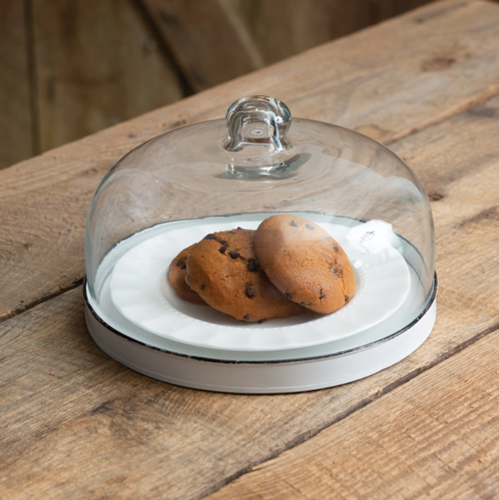 A white dessert cloche with a glass lid and a distressed metal base, featuring rustic black trim along the rim, containing two cookies.