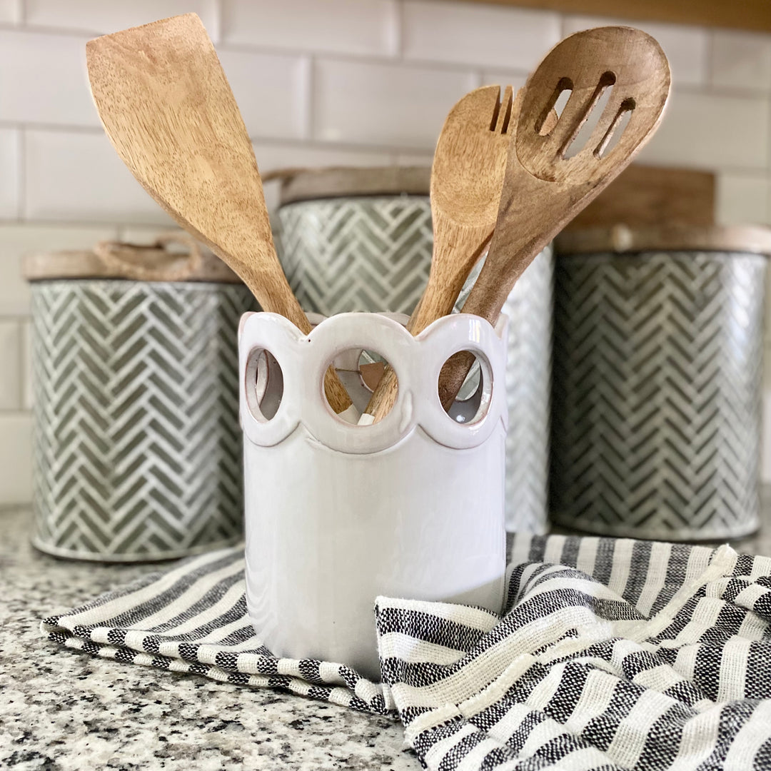 A white scalloped utensil holder made of terracotta, displayed on a kitchen countertop filled with wooden utensils, with a striped patterned dishtowel and decorative jars in the background.